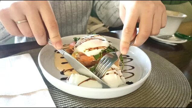 Close up of female hands using fork and knife to cut a poached egg with a soft runny yolk on toasted bread with salmon and fresh greens.