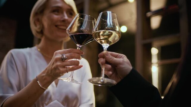 Slowmo selective focus shot of hands of elegant couple in formalwear clinking glasses with red and white wine while standing in luxury restaurant