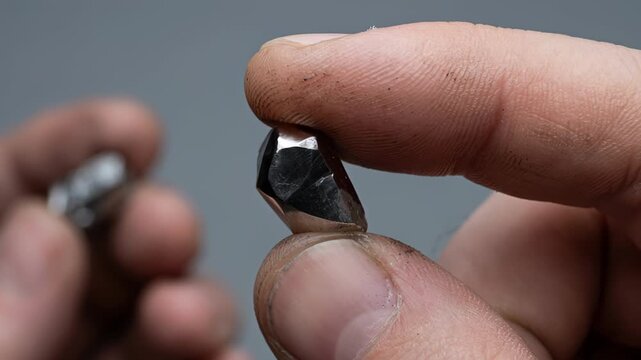 Miner's dirty hand holding a small, rough pyrite mineral, rotating the raw gemstone to inspect its metallic luster and crystal structure against a blurred background, showcasing the valuable ore