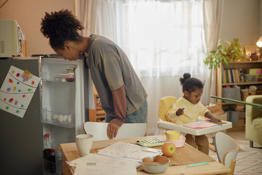 Black teenage girl checking nearly empty refrigerator while child drawing at table, showing household financial hardship. Ideal for poverty, food insecurity, budgeting, social support campaigns