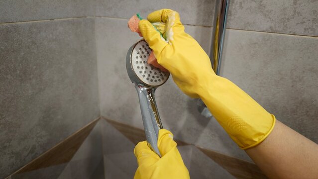 Person cleans a shower head with a sponge and wears yellow gloves in a bathroom