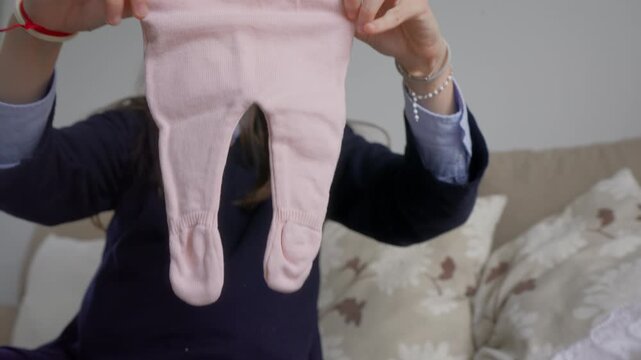 Woman hands holding up small pink knitted baby trousers, slow motion close up, stack of folded baby clothes in background, soft indoor light