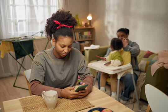 Teenage Black girl checking smartphone at table while mother caring for child in background, showing low income family life. Useful for poverty, financial stress, social support campaigns