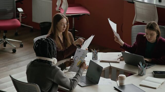 High angle shot of multiethnic team of gen Z colleagues exchanging business documents and analyzing data while collaborating at shared desk in modern office