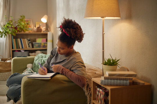 African American child studying at home, writing in notebook while sitting on sofa, showing low income family life and education access. Useful for poverty, support programs, social aid campaigns