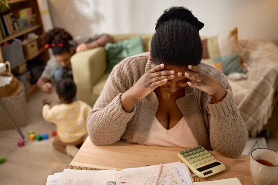 Black mother holding head while reviewing bills at home, facing financial stress as children playing nearby. Ideal for poverty, debt, family hardship, budgeting campaigns, medium shot
