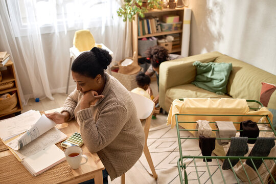 Wide angle view of African American mother reviewing household bills while caring for children at home, showing financial hardship. Ideal for poverty, budgeting, debt, welfare campaigns