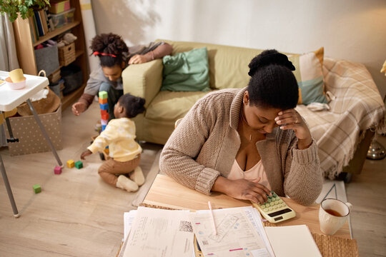 Black mother calculating household expenses while children playing in living room, showing financial stress and poverty. Useful for budgeting, debt, low income, family hardship campaigns