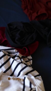 Unfolded clothes lie scattered on a surface as additional garments fall into the frame one by one, thrown by an unseen person. 