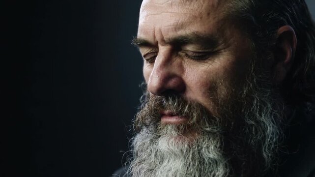 Mature man with long gray beard eyes closed against dark background, close up