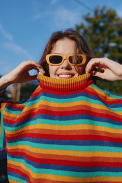 Woman smiling outdoors in a colorful striped sweater and yellow sunglasses, young adult in casual outfit and turtleneck, playful pose in park under sunny day light.
