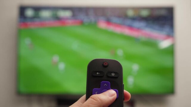 Person holding a tv remote controller and changing channels while watching a soccer match, representing modern entertainment technology and leisurely home activities