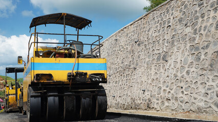 Heavy paver machine stands near a textured rock wall during road construction. Low angle emphasizes its solid industrial power under bright sunny weather. © wahid HA