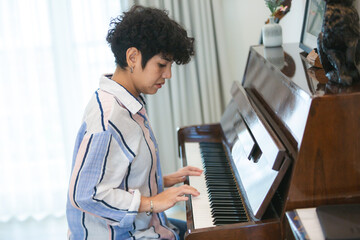 Young asian woman curly hair striped shirt casual clothing wrist bracelet upright piano home interior window curtain natural light concentration portrait of young asian woman with curly hair © chokniti