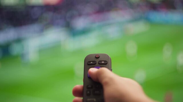 Woman's hand using a television remote control to change channels while watching a live football or soccer match on a large screen at home, creating a cinematic blur effect