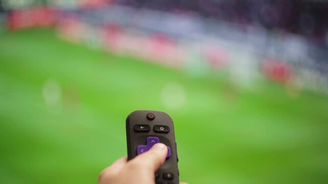 Woman's hand using a smart tv remote to change channels, surfing on demand television while watching a live football game at home on a modern big screen television