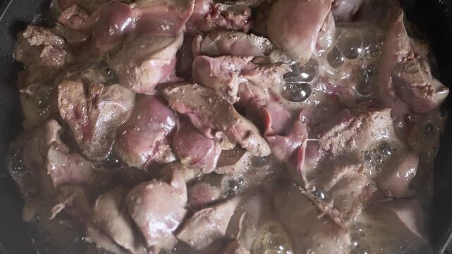 Raw chicken livers sizzle in a hot frying pan with rendered fat bubbles and intense heat texture. Concept of culinary preparation, traditional offal cooking, and home kitchen process.