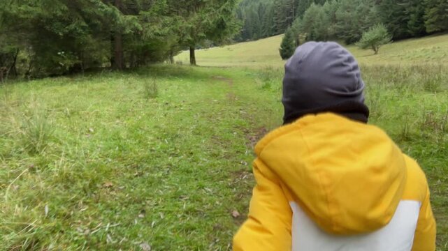 Tracking shot of a young child in a yellow jacket walking on a path, close-up back view, moving through a green meadow with a forest background, slow-motion footage