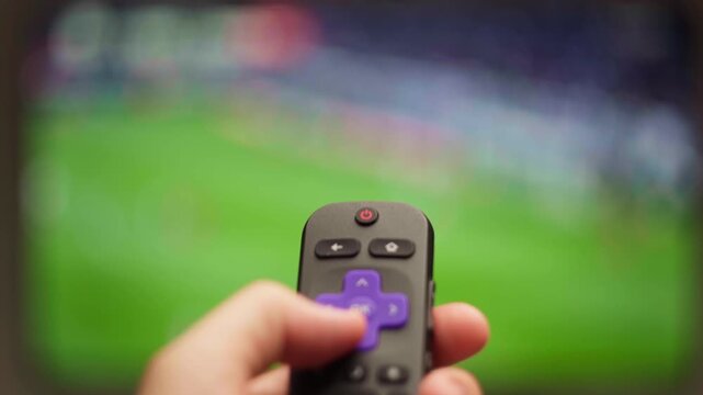 Person using a smart tv remote control for watching a live sports game on a streaming service, zapping through channels and navigating the on-screen menu