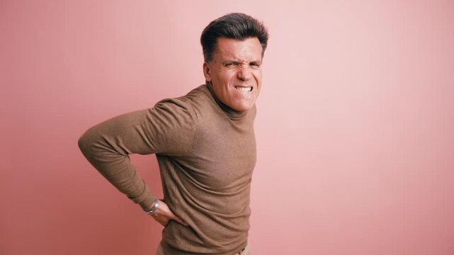 Middle aged man experiencing severe lower back pain and discomfort. He holds his aching lumbar region with a pained expression on his face