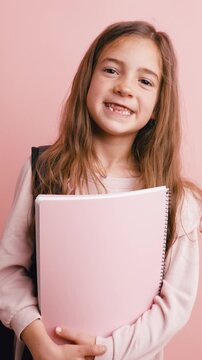 Portrait of a happy little girl with a backpack holding a pink notebook. Young pupil smiling at the camera, ready for back to school. Vertical
