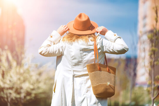 Woman in hat with straw bag walking in park. Back view of fashionably dressed traveler during warm spring day.