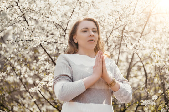 Woman praying with closed eyes in blooming garden. Meditation and mindfulness practice during peaceful spring morning.