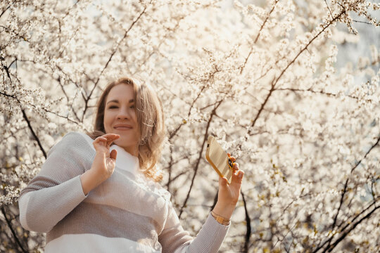 Woman waving hand during video call in blooming garden. Digital interaction and social connection in nature.