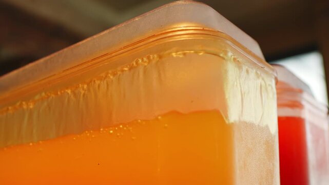 Transparent plastic dispenser filled with golden orange juice, foam layer at liquid surface and condensation on outer wall, slow motion, red container blurred behind