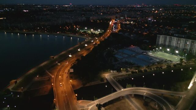 Aerial night drone shot of the illuminated Ciurel bridge in Bucharest, Romania. View of the cable-stayed bridge with traffic light trails and city lights reflecting in the Dambovita river.
