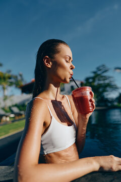 Candid outdoor moment of a woman in a white bikini beside a pool, sipping a refreshing drink through a straw, sunlit skin, relaxed posture, vibrant colors, summer lifestyle by the water