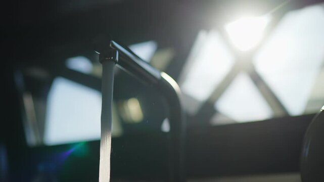 Stream of water pours from modern metal faucet against blurred bright window, slow motion, lens flare and geometric light pattern visible in background