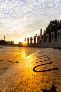 View of the World War II Memorial at sunrise with a sunburst effect over the stone pillars and the Washington Monument in the distance in Washington, District of Columbia, United States.