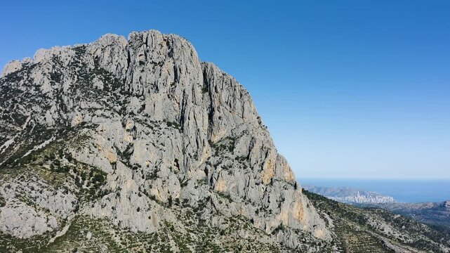 Aerial view of the Puig Campana mountain range and Benidorm city far away, characterized by its vertical mountain-shaped walls and its different vegetation between the shaded area and the sunny area.