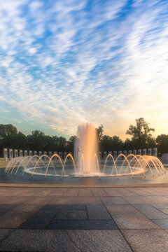 View of the World War II Memorial fountain with illuminated water jets and stone columns under a wispy sky at sunset in Washington, District of Columbia, United States.