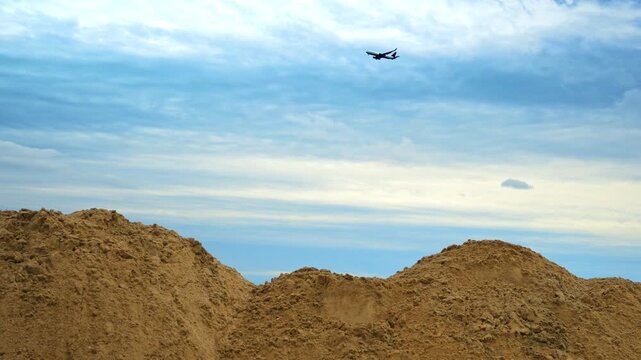 Plane flying rather low in the overcast sky. Low angle view. Piles of yellow sand in the foreground.