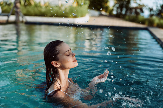 Relaxed woman enjoys a quiet moment in a sunlit pool, eyes closed, feeling water droplets and a cool breeze as she rises above the surface, portraying a calm outdoor lifestyle, leisure, and