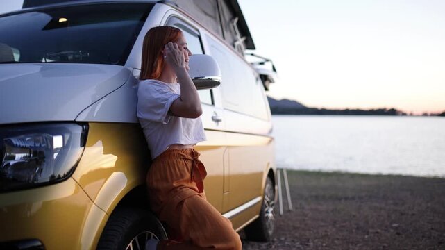 Pensive young woman with red hair leaning against her camper van. She is enjoying the beautiful sunset view by a peaceful lake on her road trip