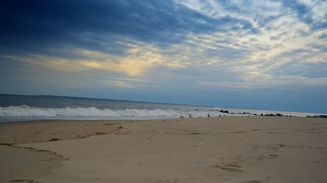 Clouds covered the sky over the raging sea. Seagulls walk by the beach in the backdrop.