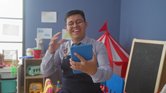 Man teacher holding blue tablet and smiling in school building, wearing glasses and apron straps, toys and red play tent in kindergarten room; joy.