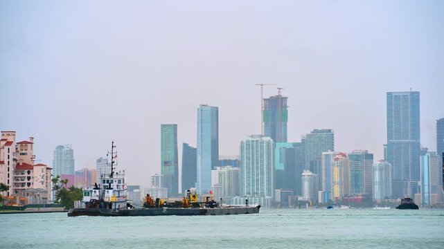 Industrial tugboat barge with Miami downtown skyline background. Work barge and tugboat on water with modern city buildings and construction cranes under overcast sky.