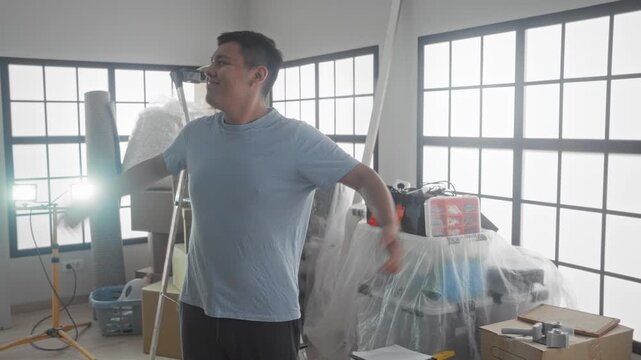 Man standing with hands on hips beside covered furniture, stacked moving boxes, ladder and tool case in a building during move in; pride.