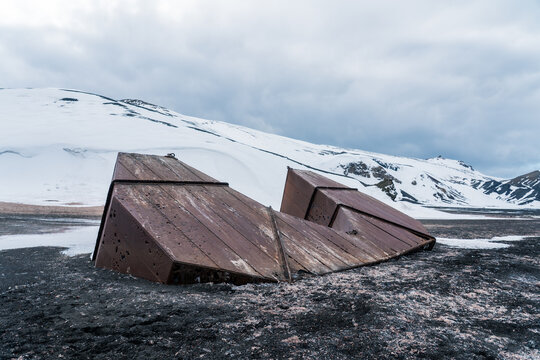 View of the rusted iron remains of an abandoned whaling station on the dark volcanic sand of Deception Island with snow-covered hills Antarctica.