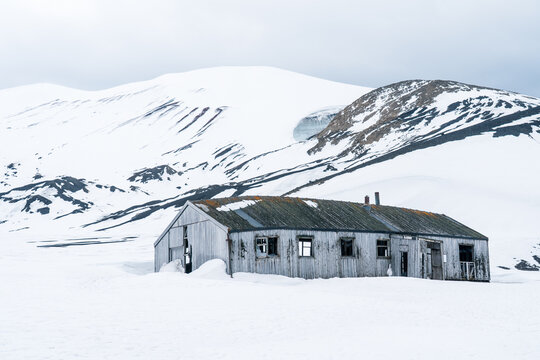 View of an abandoned wooden building at Deception Island with snow-covered mountains and an overcast sky Antarctica.