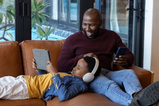 African American father and son relaxing on tan leather sofa at home using tablet, white headphones