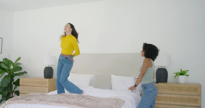 Diverse female friends led by woman in yellow top jumping, dancing on bed in bedroom playfully
