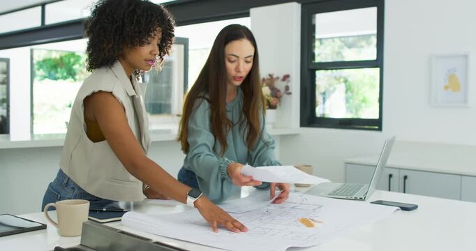 Diverse female coworkers starting design review, unrolling plans on kitchen island and using laptop