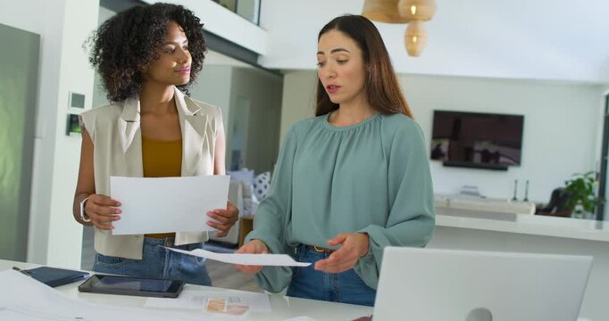 Diverse female coworkers holding sheets, reviewing blueprint at counter, sliding swatches, planning