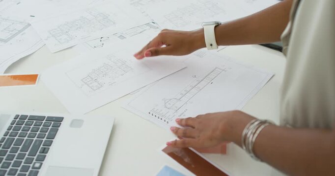 Adult female smoothing and aligning printed design plans at desk for review with laptop and tablet