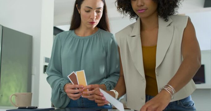 Two women standing at home counter left holding 3 swatches sorting samples near tablet for design